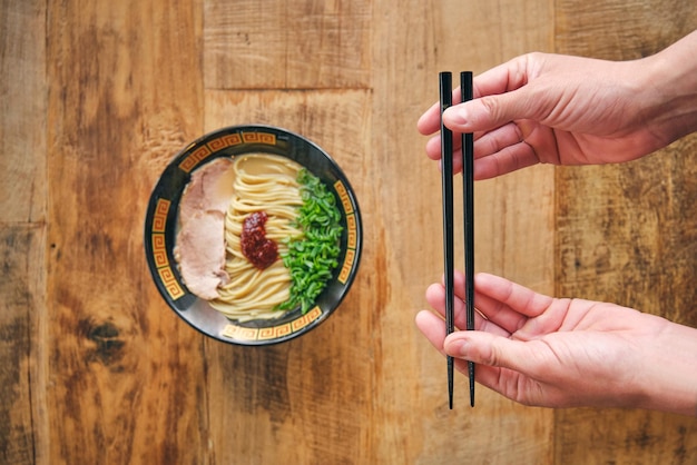 Overhead view of crop anonymous person with food sticks over yummy Japanese noodle soup in bowl on wooden table