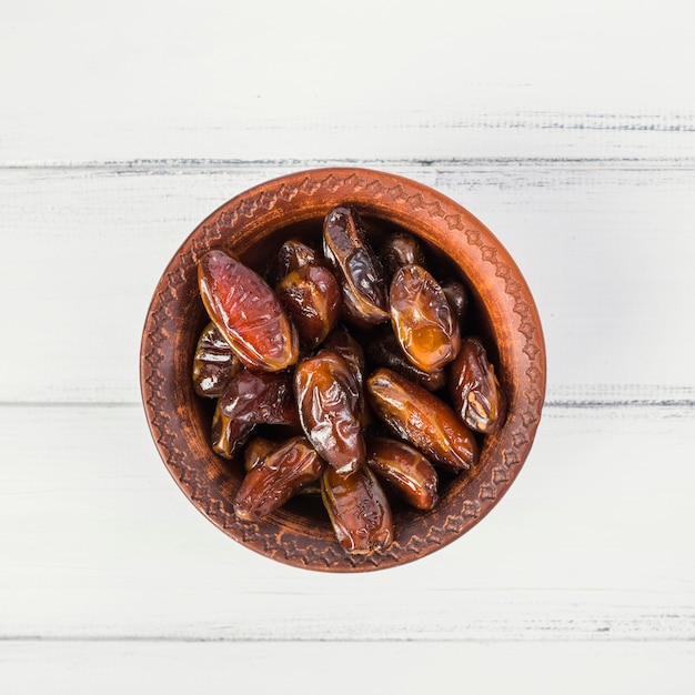 An overhead view of dates in a wooden bowl on white wooden table