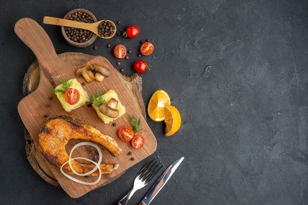 Overhead view of delicious fried fish and mushrooms tomatoes greens on wooden cutting board cutlery set pepper on black surface