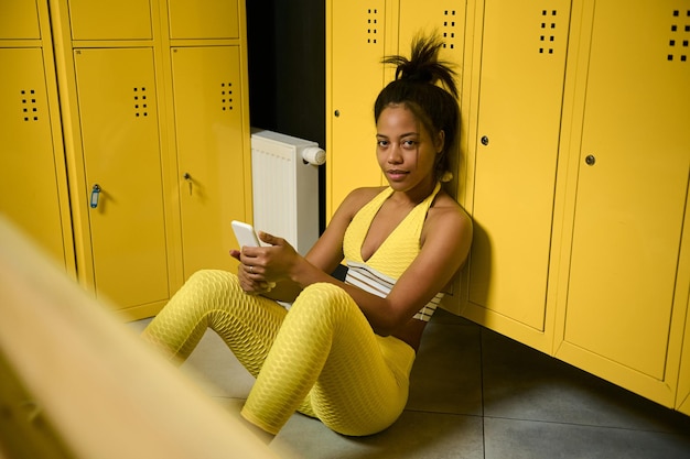 Overhead view of a female African athlete, sportswoman in yellow tight-fitting tracksuit, resting after heavy workout, sitting on the floor in gym locker room, swiping in smartphone