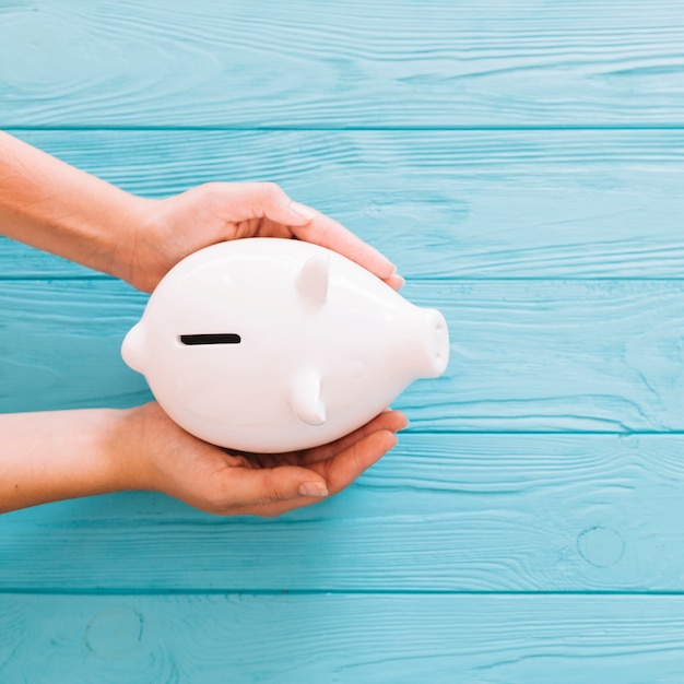 Photo overhead view of hand protecting white piggybank on wooden blue background