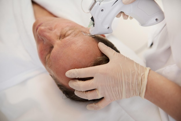 Overhead view of a handsome aged man lying on massage table during mesotherapy gun therapy in modern spa clinic. Anti-aging, rejuvenation and alternative beauty treatment to prevent first wrinkles