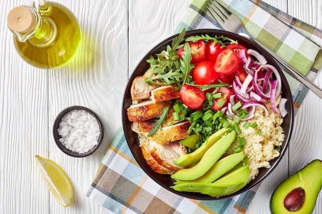 Overhead view of healthy chicken bowl with steamed couscous, fresh vegetables, avocado, scallion and arugula on a white wooden table with ingredients, horizontal view from above, close-up