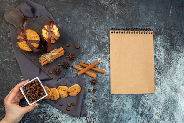 Overhead view of homemade cookies cinnamon limes and a cup of tea on wooden cutting board on dark color towel hand holding beans notebook on ice background