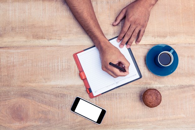 Punto di vista ambientale dell'uomo che scrive sul diario al tavolo da caffè e torta della tazza