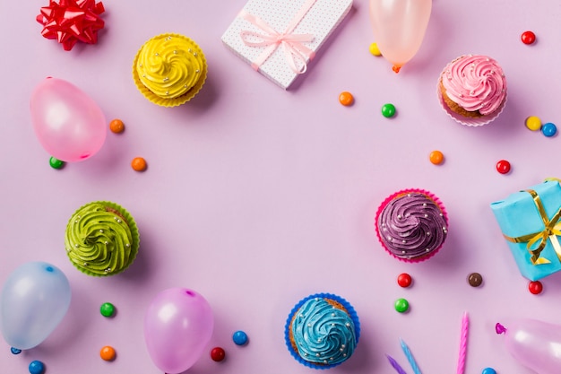 Una vista dall'alto di muffin; palloncini; gemme; confezione regalo e candele su sfondo rosa