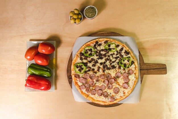 Overhead view of a pizza with hot dogs and vegetables and surrounding decorations on a wooden table.