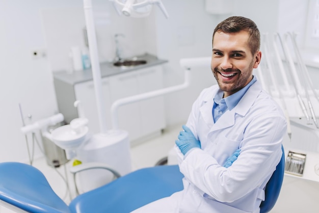 Вид сверху улыбающегося врача-мужчины со скрещенными руками, смотрящего в камеру в стоматологической клинике