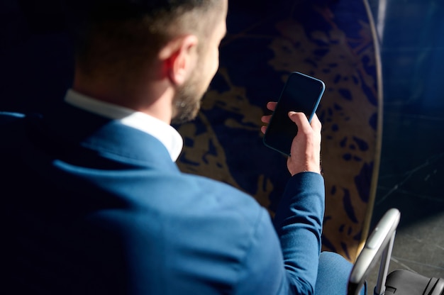 Overhead view of a well-groomed attractive handsome bearded businessman in business casual suit using a mobile phone while sitting on a chair in a hotel lobby during his work trip. Copy space