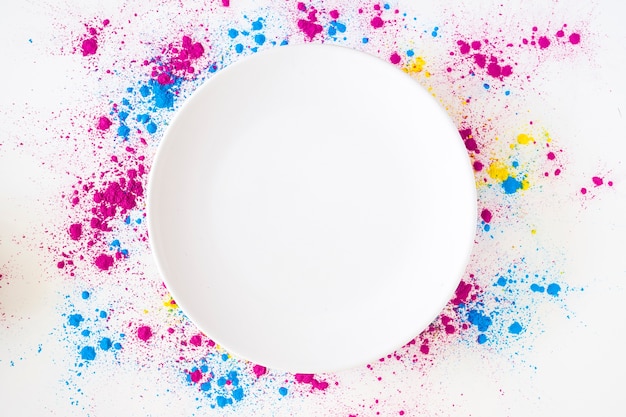 An overhead view of white plate on holi color powder over white background