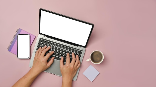 Overhead view of woman hands typing on laptop computer with smart phone and coffee cup on pink background