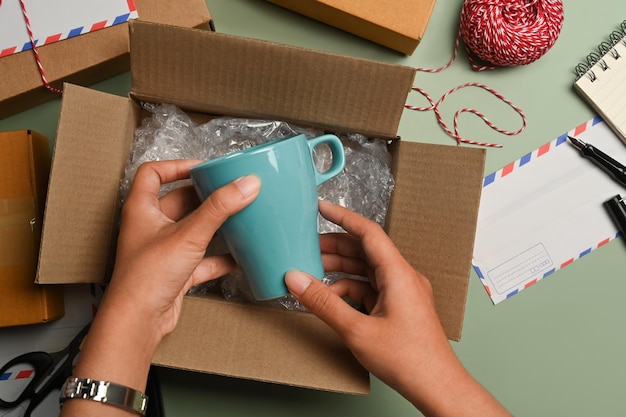 Overhead view of woman putting ceramic cup into cardboard box preparing to pack gifts for holiday season