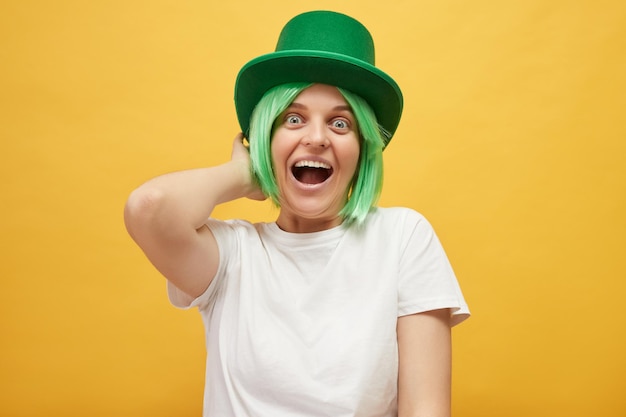 Foto overijdelijke aantrekkelijke vrouw met groen haar met een leprechaun hoed die geïsoleerd staat op een gele achtergrond en schreeuwt met een vrolijk gezicht en feest viert