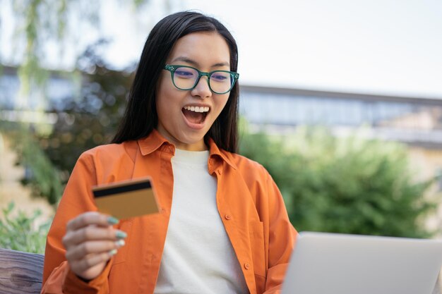 Foto donna asiatica felicissima di affari che tiene la carta di credito facendo uso del computer portatile che compera online