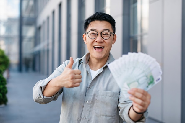 Overjoyed asian businessman holding fan of euro money and showing thumb up gesture standing outdoors