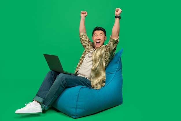 Overjoyed asian man making yes gesture sitting in beanbag chair
with laptop celebrating success over