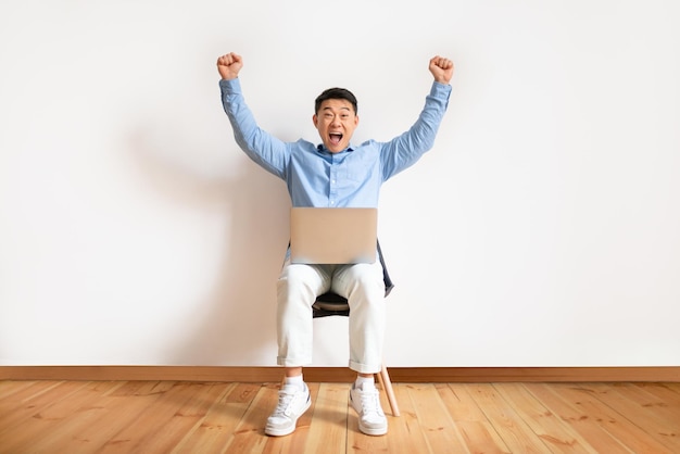 Overjoyed asian man making YES gesture sitting on chair with laptop celebrating success against white studio wall