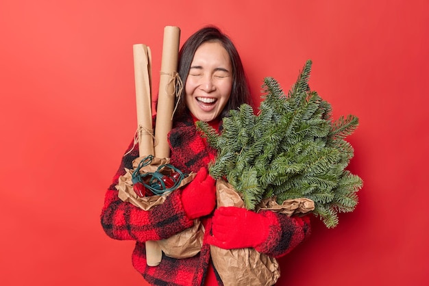 Overjoyed Asian woman with dark hair laughs out positively embraces bouquet of spruce branches rolled paper for decor and retro garland isolated over vivid red background Celebration concept