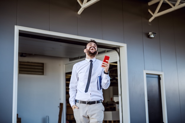 Overjoyed bearded Caucasian CEO in shirt and tie standing in front of printing shop and using smart phone.