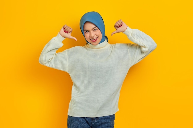 Overjoyed beautiful Asian woman in white sweater pointing thumbs at herself with proud isolated over yellow background