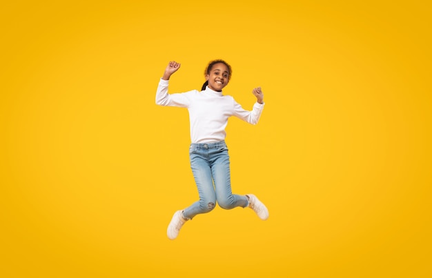 Фото Обрадованная черная маленькая девочка прыгает, весело позирует в воздухе и улыбается в камеру на желтом фоне студии