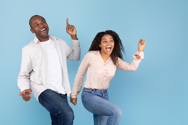 Overjoyed black middle aged man and young woman having fun together and dancing on blue background copy space