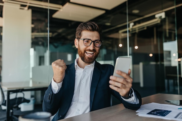 Overjoyed businessman in suit looking at smartphone and clenching fists making victory gesture in office interior