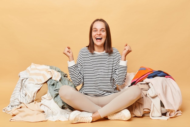 Photo overjoyed caucasian woman posing near heap of multicolored unsorted clothes isolated over beige background clenched fists rejoicing buying lots new attires