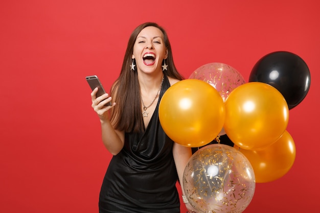 Обрадованная жизнерадостная женщина в маленьком черном платье держит воздушные шары с помощью мобильного телефона во время празднования изолированного на красном фоне. День святого Валентина, концепция праздника макета дня рождения с новым годом.