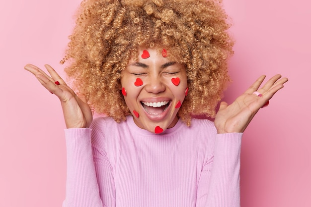 Overjoyed curly haired woman exclaims gladfully keeps palms raised laughs positively wears casual junmper has face decorated with small red hearts enjoys Valentines Day poses against pink wall