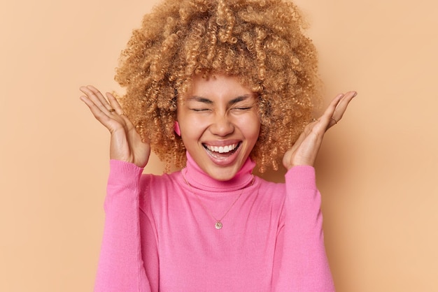 La donna dai capelli ricci felicissima alza i palmi vicino alla testa esclama ad alta voce dalla felicità tiene gli occhi chiusi vestita in abiti casual reagisce alle buone notizie isolate su sfondo beige. emozioni umane