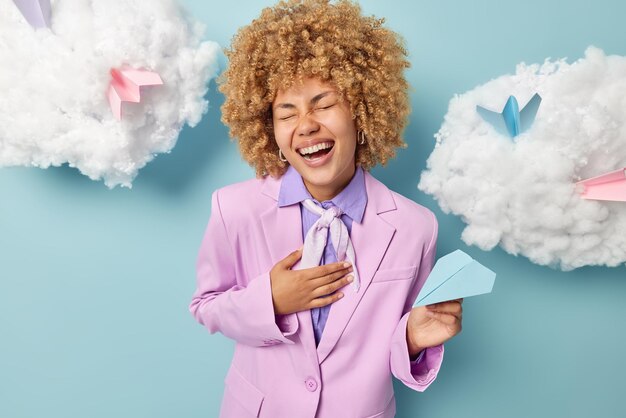 Overjoyed curly woman keeps hand on chest cannot stop laughing\
dressed in formal clothig prepares for business meeting holds\
handmade paper plane isolated over blue background white clouds\
above