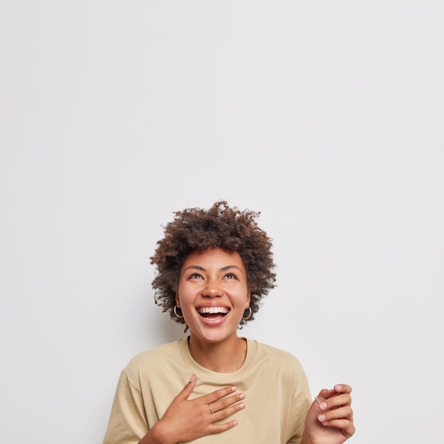 Overjoyed curly woman laughs joyfully keeps hand on chest concentrated above concentrated overhead wears casual beige t shirt dressed casually isolated over white background with blank copy space