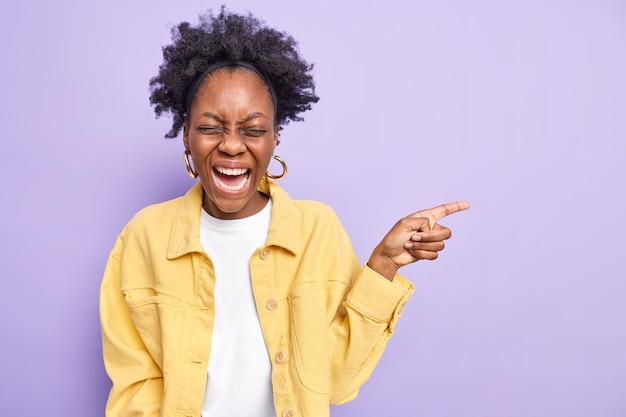 Overjoyed dark skinned woman with curly hiar points away on copy space