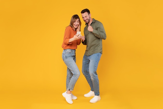 Overjoyed european spouses holding smartphone and shaking fists in joy and excitement reading great