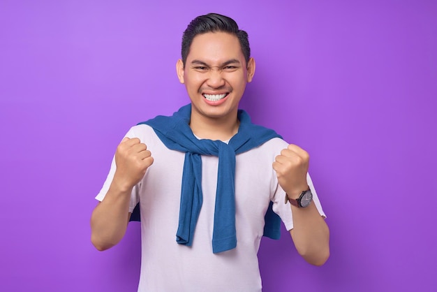 Overjoyed and excited young asian man 20s wearing white tshirt doing a winner gesture celebrate clenching fists say yes isolated on purple background people lifestyle concept