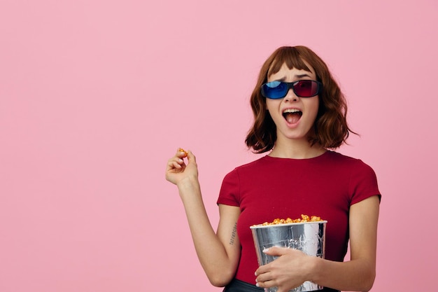 Overjoyed happy cute redhead lady in red tshirt 3D glasses with popcorn watch movie posing isolated on over pink studio background Copy space Banner Fashion Cinema concept Entertainment offer