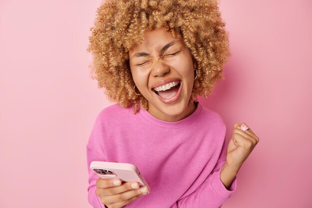 Overjoyed happy european woman gestures yes glad to win mobile\
game celebrates great news laughs joyfully dressed in casual\
pullover enjoys victroy celebration isolated over pink\
background