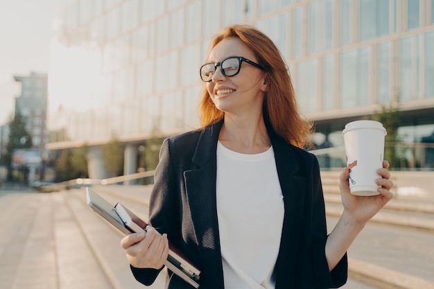 Обрадованная счастливая рыжая женщина в очках пьет кофе и держит ноутбук во время прогулки на свежем воздухе