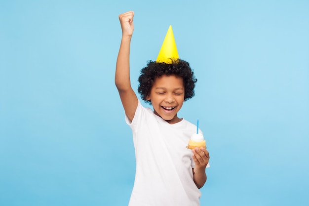 Photo overjoyed happy little boy with party cone holding cupcake and keeping eyes closed while making wish on his birthday standing with raised hand shouting from happiness studio shot blue background