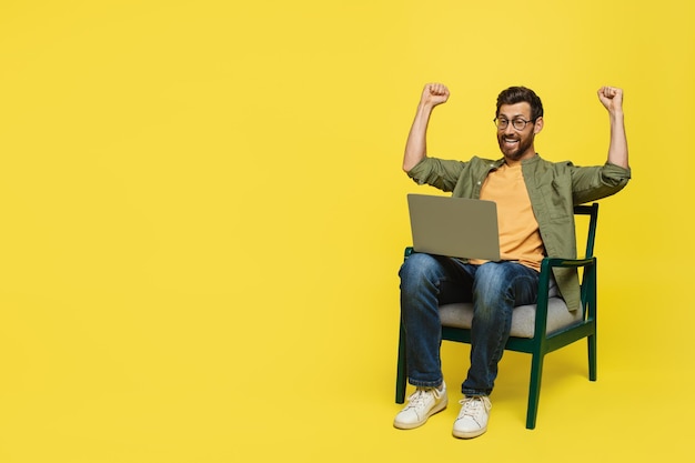 Overjoyed man sitting on chair with laptop and shaking fists celebrating success yellow background copy space