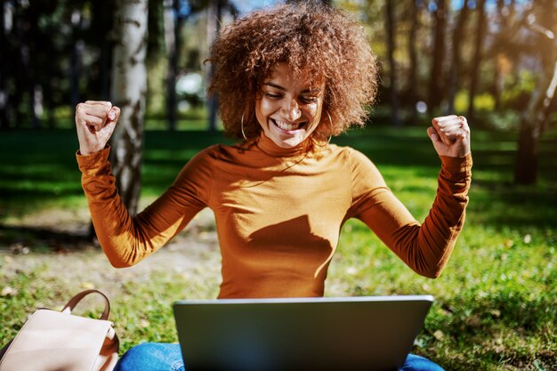 Overjoyed pretty young mixed race woman dressed casual and with curly hair cheering for success and looking at laptop while sitting on the lawn in park.
