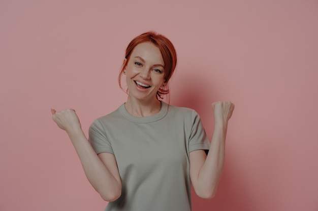 Overjoyed redhaired woman celebrating victory doing winner gesture on pink background