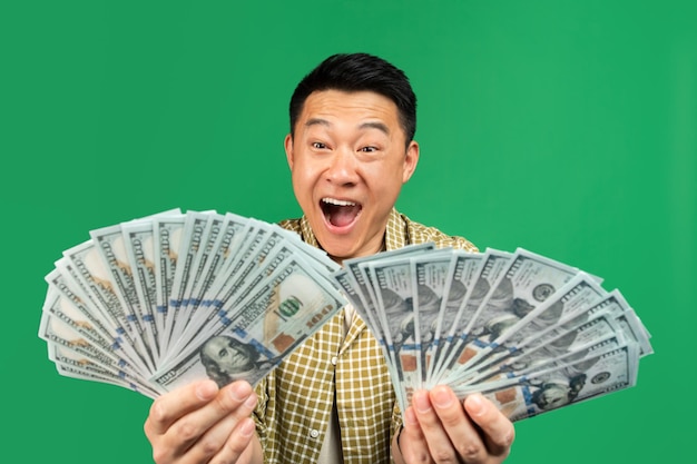 Overjoyed rich mature asian man holding two fans of money\
celebrating big financial luck over green