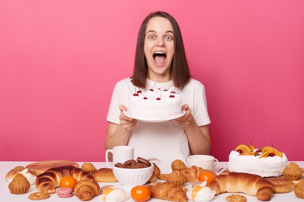 Overjoyed screaming brown haired woman in white t shirt sitting at table showing tasty cake breaking diet or having cheat meal isolated over pink background