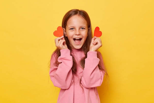 Overjoyed screaming extremely happy little girl wearing pink sweatshirt holding red little hearts isolated over yellow background yelling with happy face