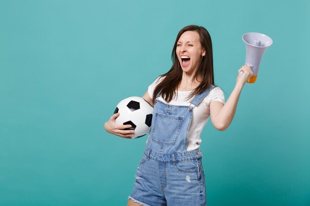 Радостно кричащая молодая женщина-футболистка поддерживает любимую команду с футбольным мячом, мегафон изолирован на синем бирюзовом фоне. Эмоции людей, концепция спортивного семейного отдыха. Скопируйте пространство для копирования.