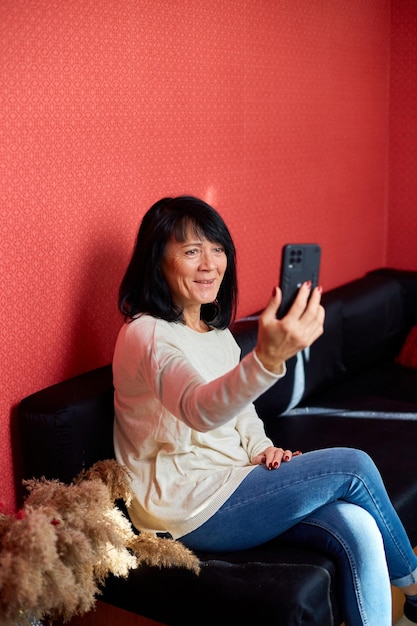Una donna anziana felicissima si siede sul divano a casa e parla in videochiamata su un cellulare moderno