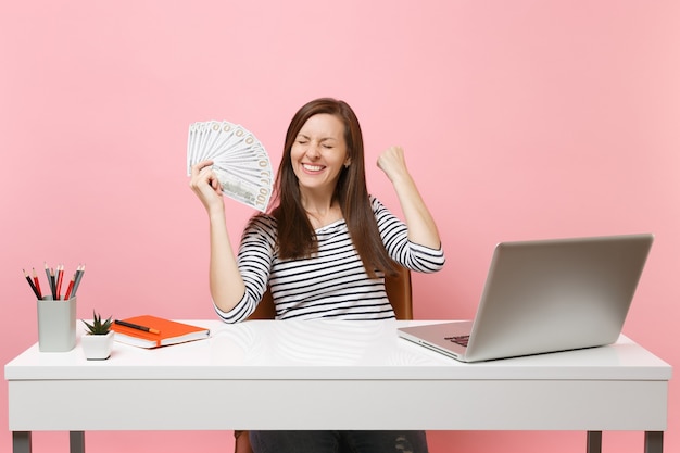 Donna felicissima che stringe i pugni come un vincitore che tiene in mano un sacco di dollari, denaro contante lavora in ufficio alla scrivania bianca con pc portatile