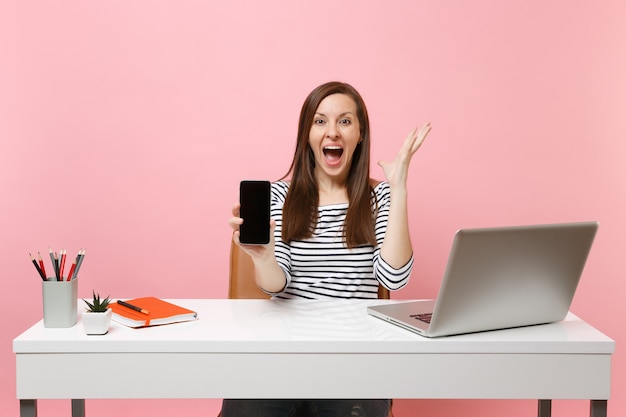 Overjoyed woman spreading hands holding mobile phone with blank empty screen work at white desk with contemporary pc laptop isolated on pastel pink background. Achievement business career. Copy space.